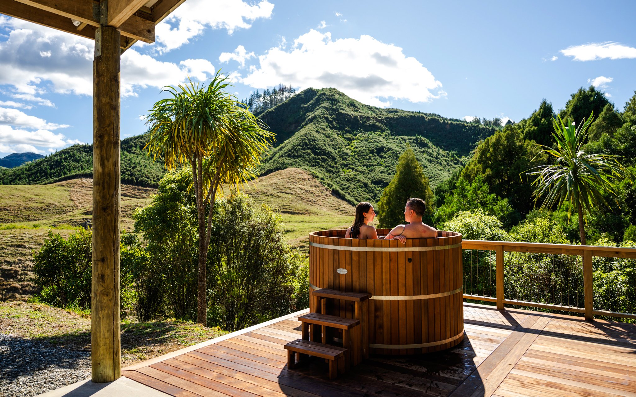 Couple relaxing in a wooden hot tub with scenic mountain views at Waitomo, New Zealand.