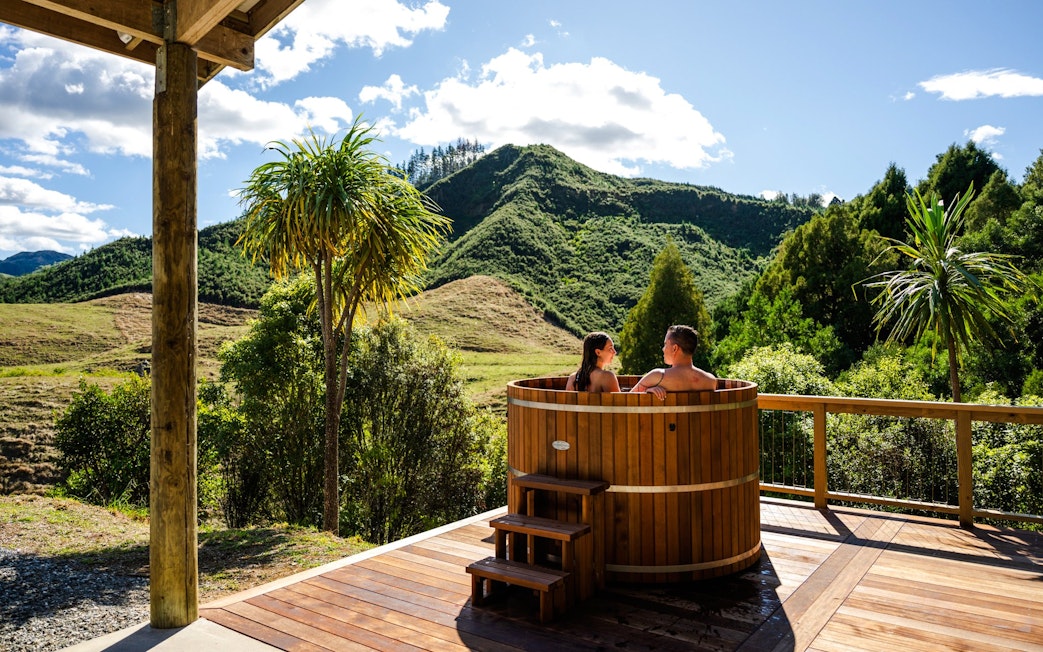 Couple relaxing in a wooden hot tub with scenic mountain views at Waitomo, New Zealand.