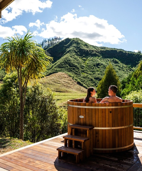 Couple relaxing in a wooden hot tub with scenic mountain views at Waitomo, New Zealand.