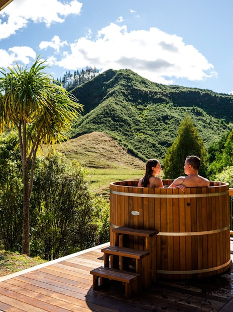 Couple relaxing in a wooden hot tub with scenic mountain views at Waitomo, New Zealand.