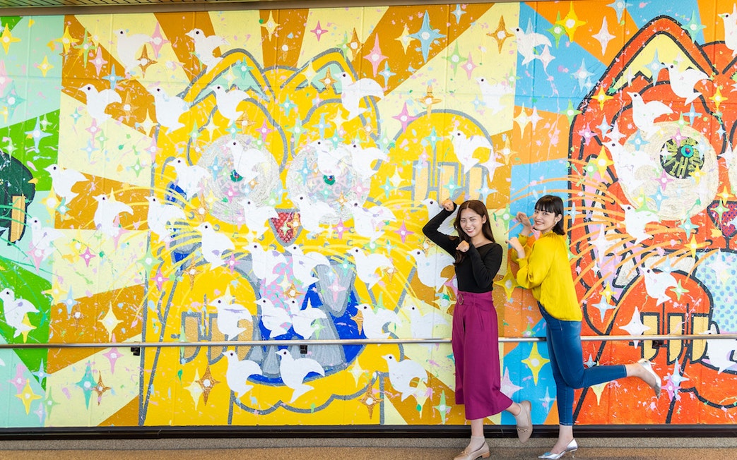 Colorful mural with doves at Hiroshima Orizuru Tower, featuring two people posing playfully.
