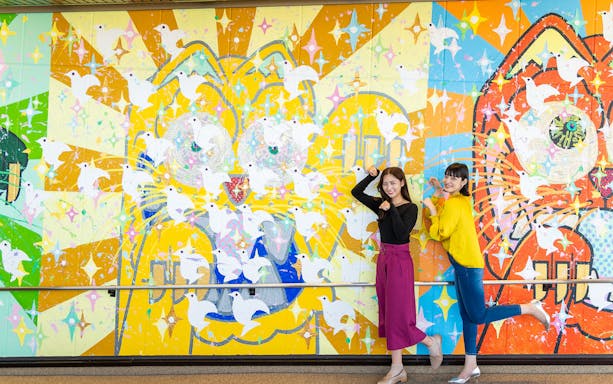Colorful mural with doves at Hiroshima Orizuru Tower, featuring two people posing playfully.