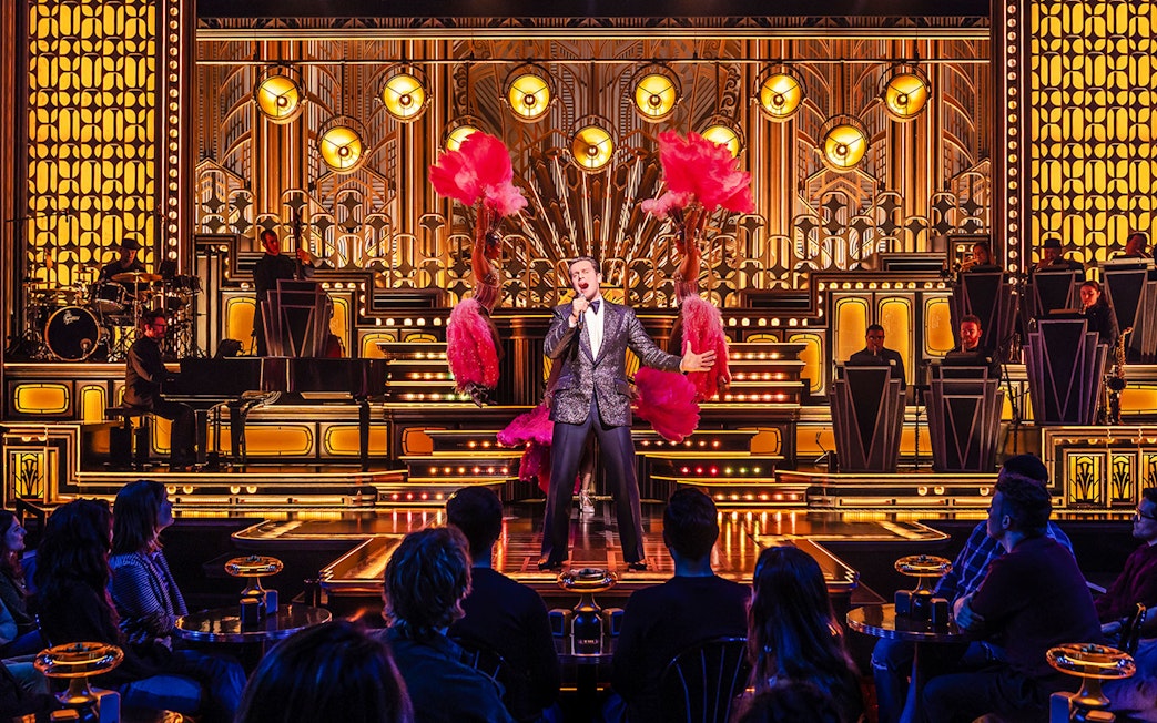 Performer singing on stage with dancers and band in a vibrant theater setting.