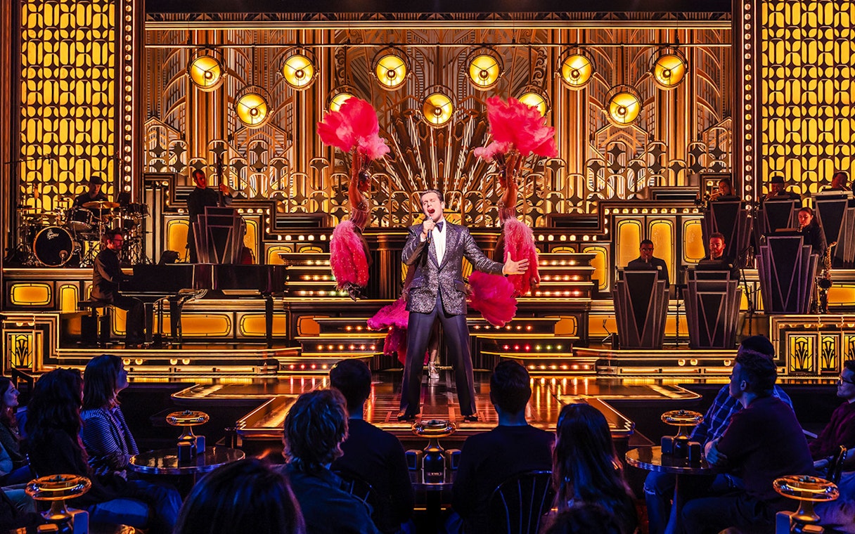 Performer singing on stage with dancers and band in a vibrant theater setting.