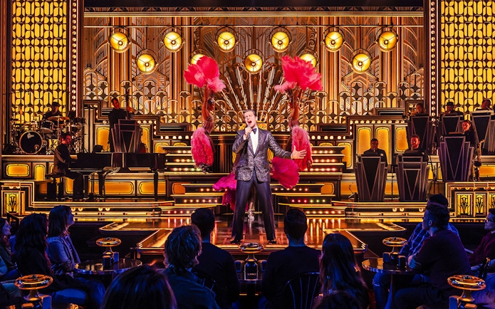 Performer singing on stage with dancers and band in a vibrant theater setting.