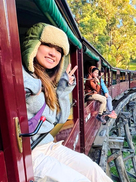 Passenger gives peace sign from Puffing Billy train window on bridge in lush forest.