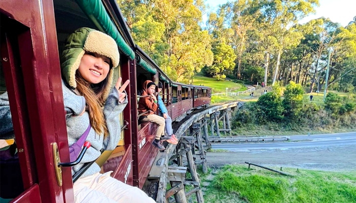 Passenger gives peace sign from Puffing Billy train window on bridge in lush forest.