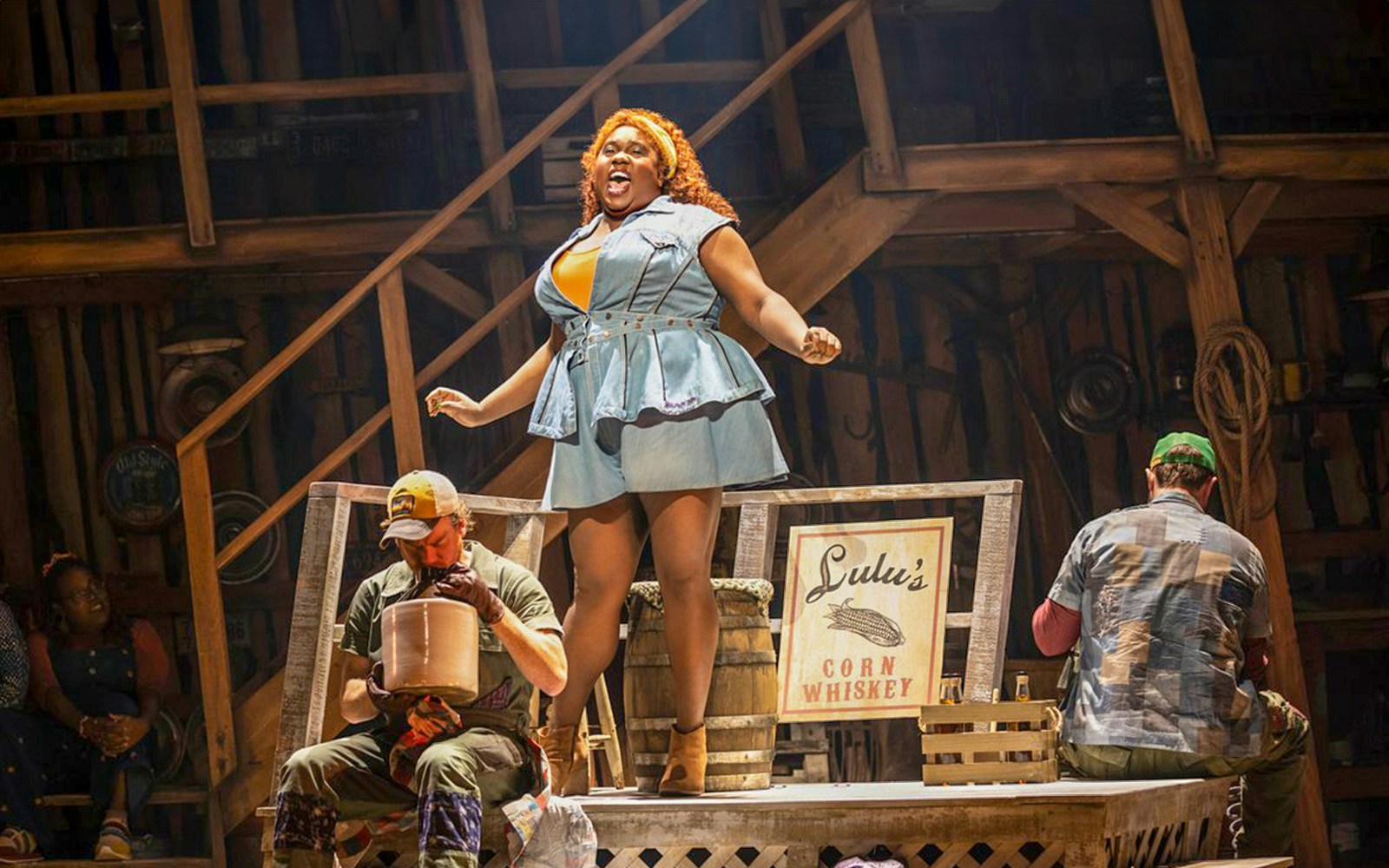 Performer singing on stage with a sign for Lulu's Corn Whiskey, related to "Shucked" show.