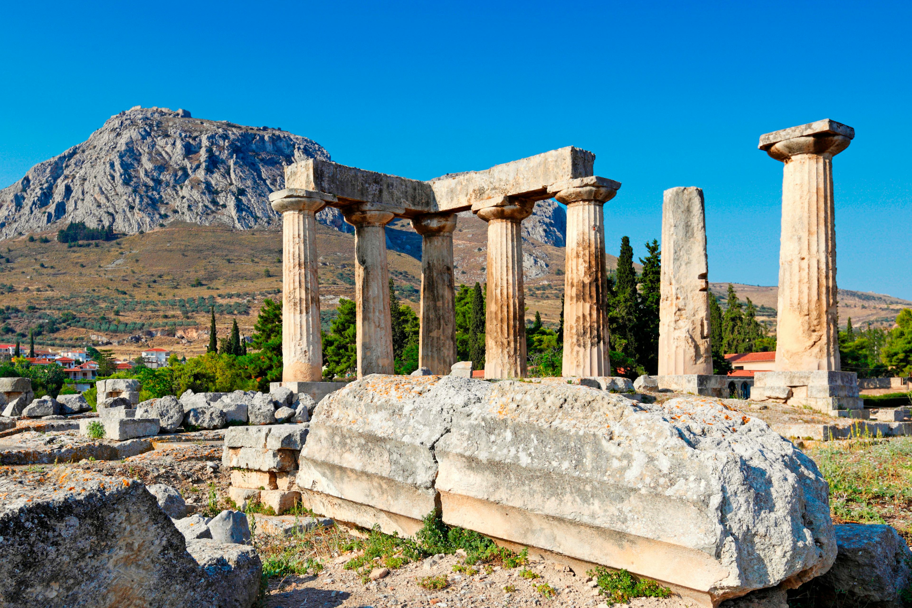 Ancient columns of the Temple of Apollo in Corinth, Greece, with a mountain backdrop.