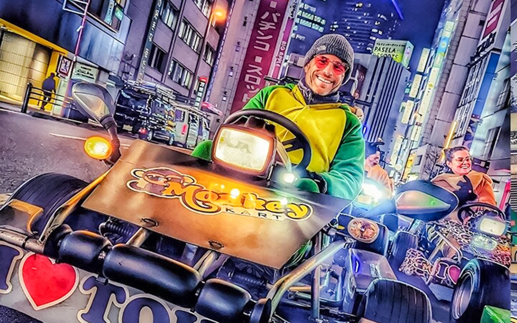 Man in green and yellow costume driving a go-kart at night in Tokyo with city lights.