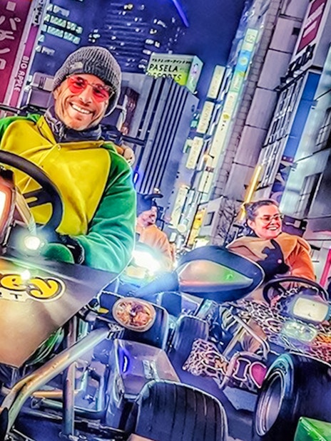 Man in green and yellow costume driving a go-kart at night in Tokyo with city lights.