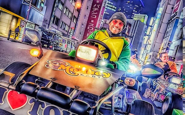 Man in green and yellow costume driving a go-kart at night in Tokyo with city lights.