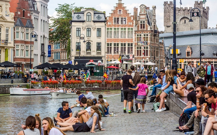People relaxing by the canals in Korenlei, Ghent, Belgium, with historic buildings in the background.