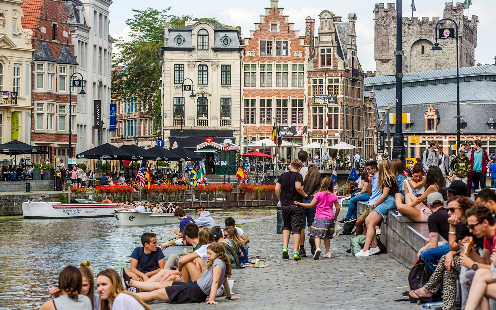 People relaxing by the canals in Korenlei, Ghent, Belgium, with historic buildings in the background.