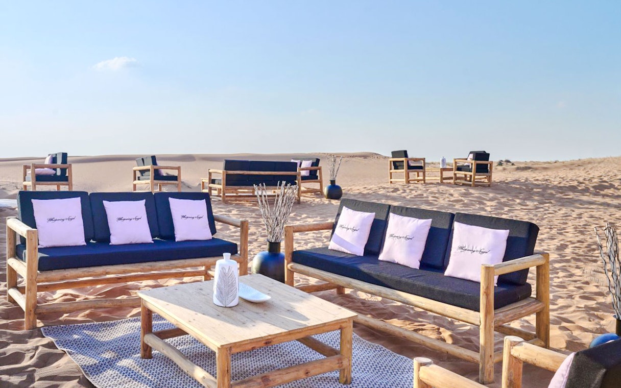 Outdoor seating at The Nomad Garden desert dining experience with wooden furniture and sand dunes.
