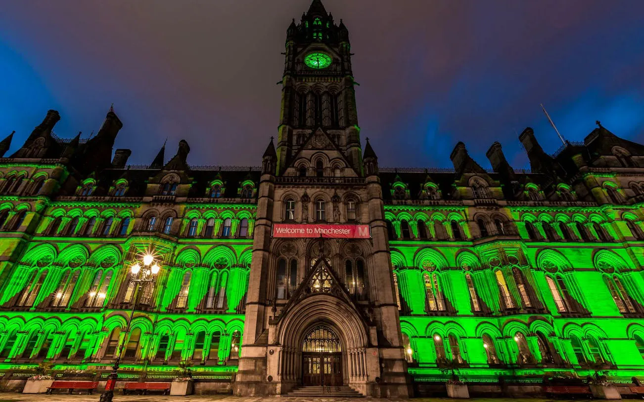 Manchester Town Hall illuminated in green at night, featuring a "Welcome to Manchester" banner.