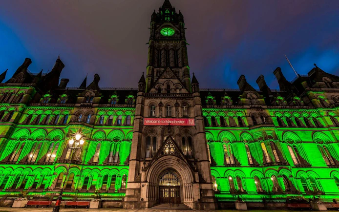 Manchester Town Hall illuminated in green at night, featuring a "Welcome to Manchester" banner.