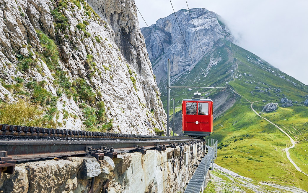 Red cogwheel train ascending Mount Pilatus, Switzerland, with rocky cliffs and green slopes.