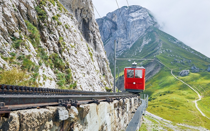 Red cogwheel train ascending Mount Pilatus, Switzerland, with rocky cliffs and green slopes.