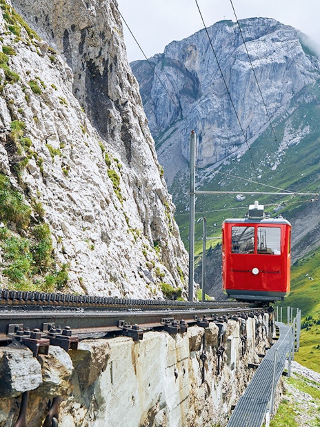 Red cogwheel train ascending Mount Pilatus, Switzerland, with rocky cliffs and green slopes.