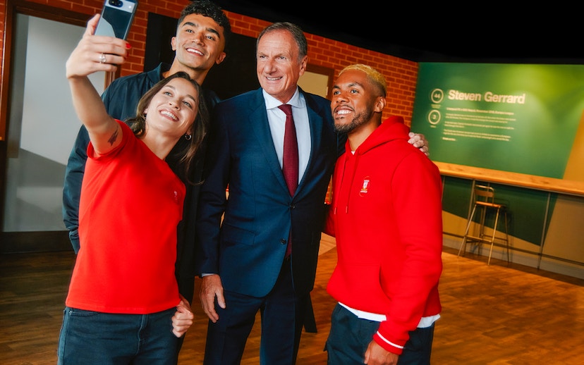 Group taking a selfie at Anfield with Liverpool Legends exhibit in background.