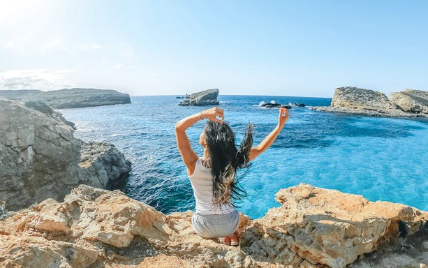 Woman sitting on rocks at The Blue Lagoon, Malta, with clear blue water in the background.