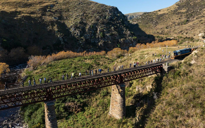 Taieri Gorge train crossing Deep Stream Bridge with tourists walking alongside in scenic landscape.