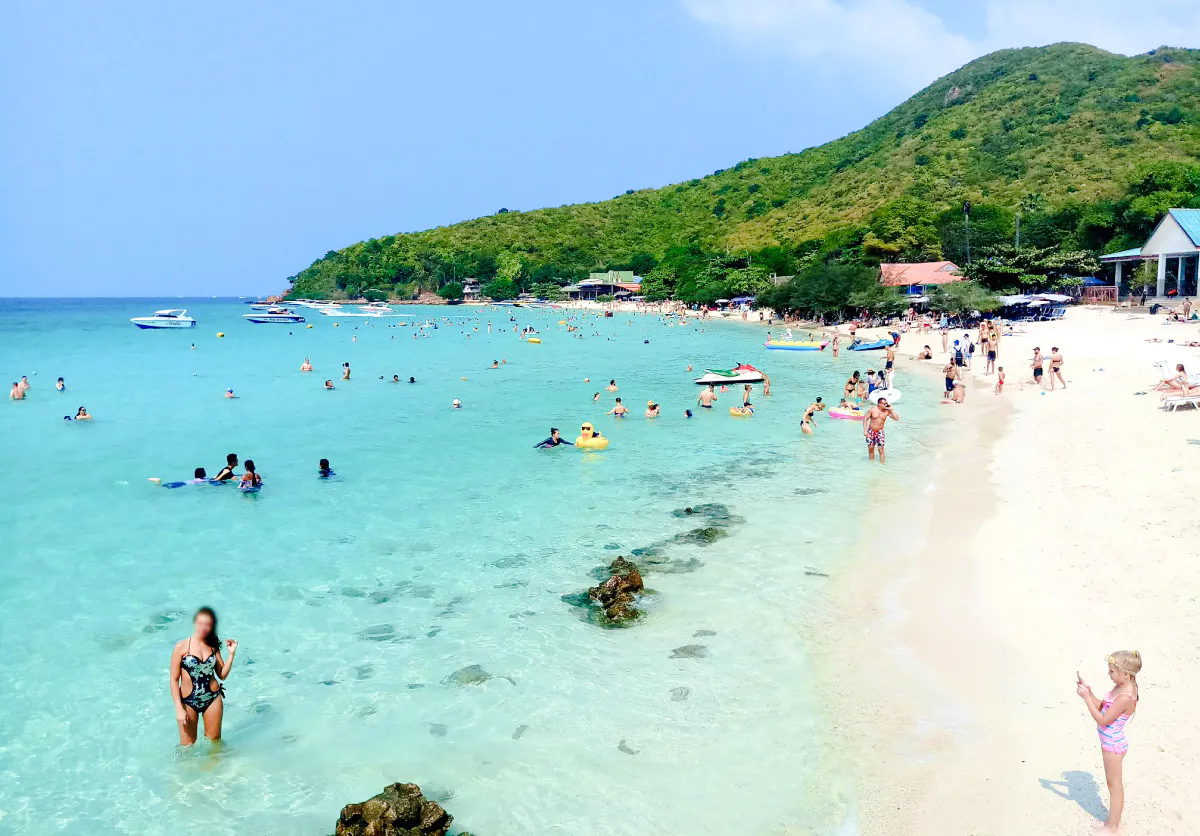 Tourists enjoying the beach and clear waters on Coral Island, Pattaya, with speedboats in the distance.