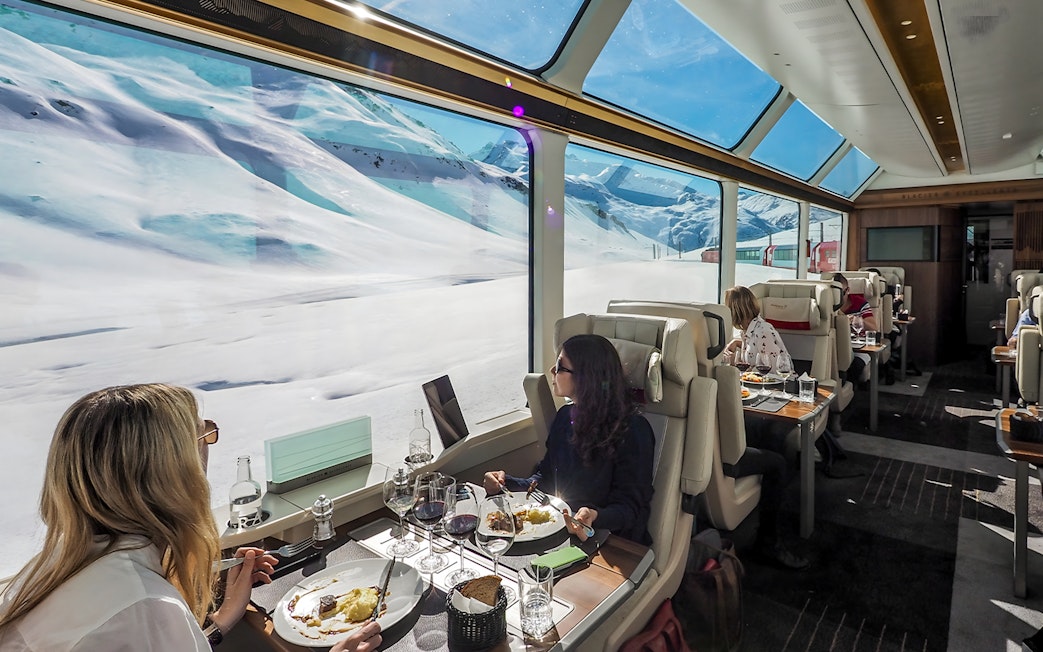 Women dining on Glacier Express with snowy mountain views.
