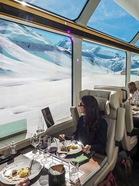 Women dining on Glacier Express with snowy mountain views.