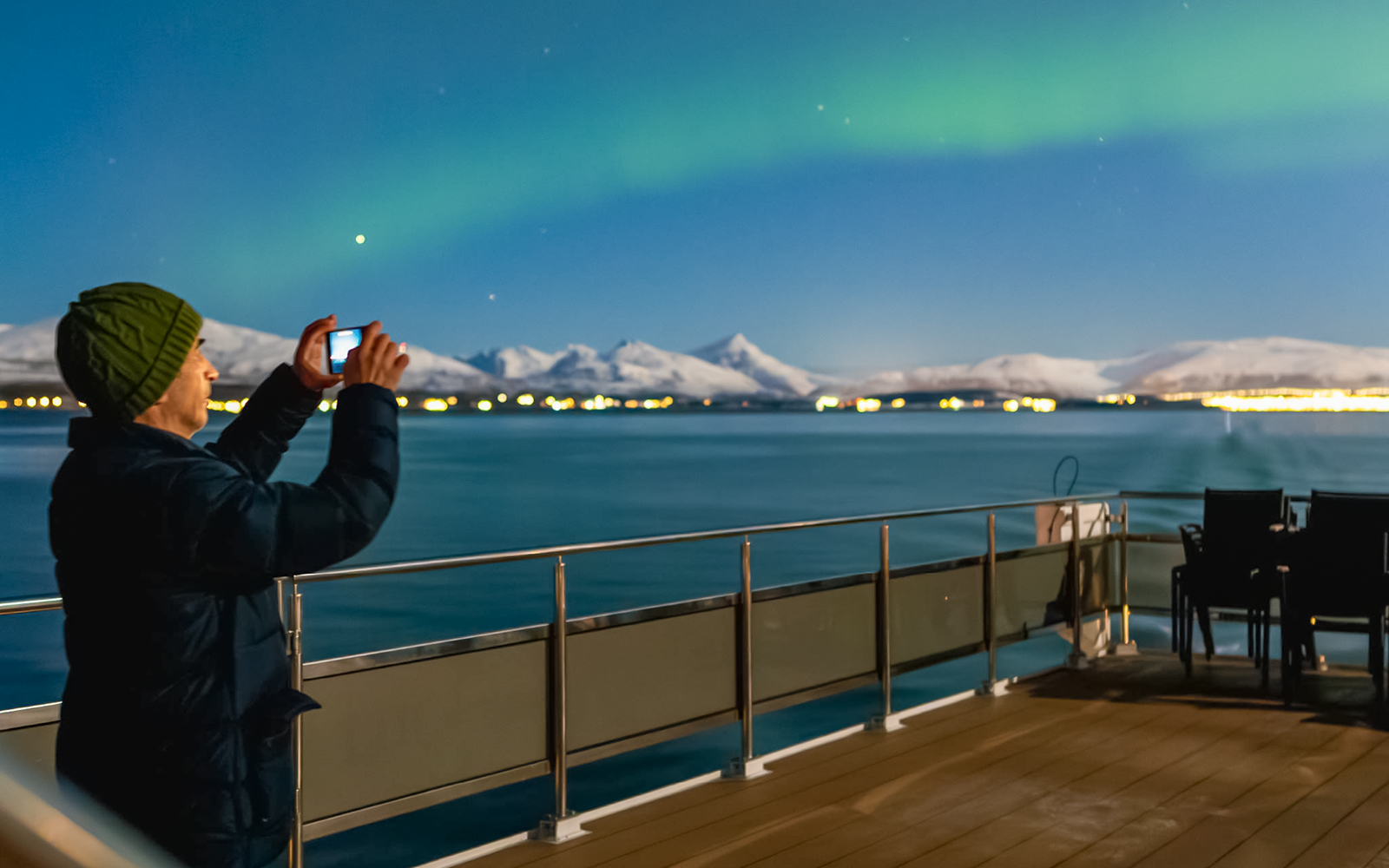 Person photographing Northern Lights from cruise deck with snowy mountains in background.