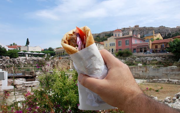 Hand holding a gyro with Athens ruins and colorful buildings in the background.