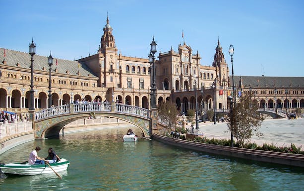 Boating on the canal at Plaza de España, Seville, with ornate bridge and historic architecture.