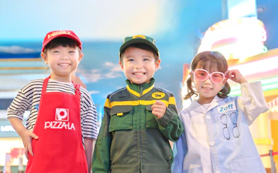 Children in themed costumes at KidZania Tokyo, including a pizza chef, delivery worker, and optician.