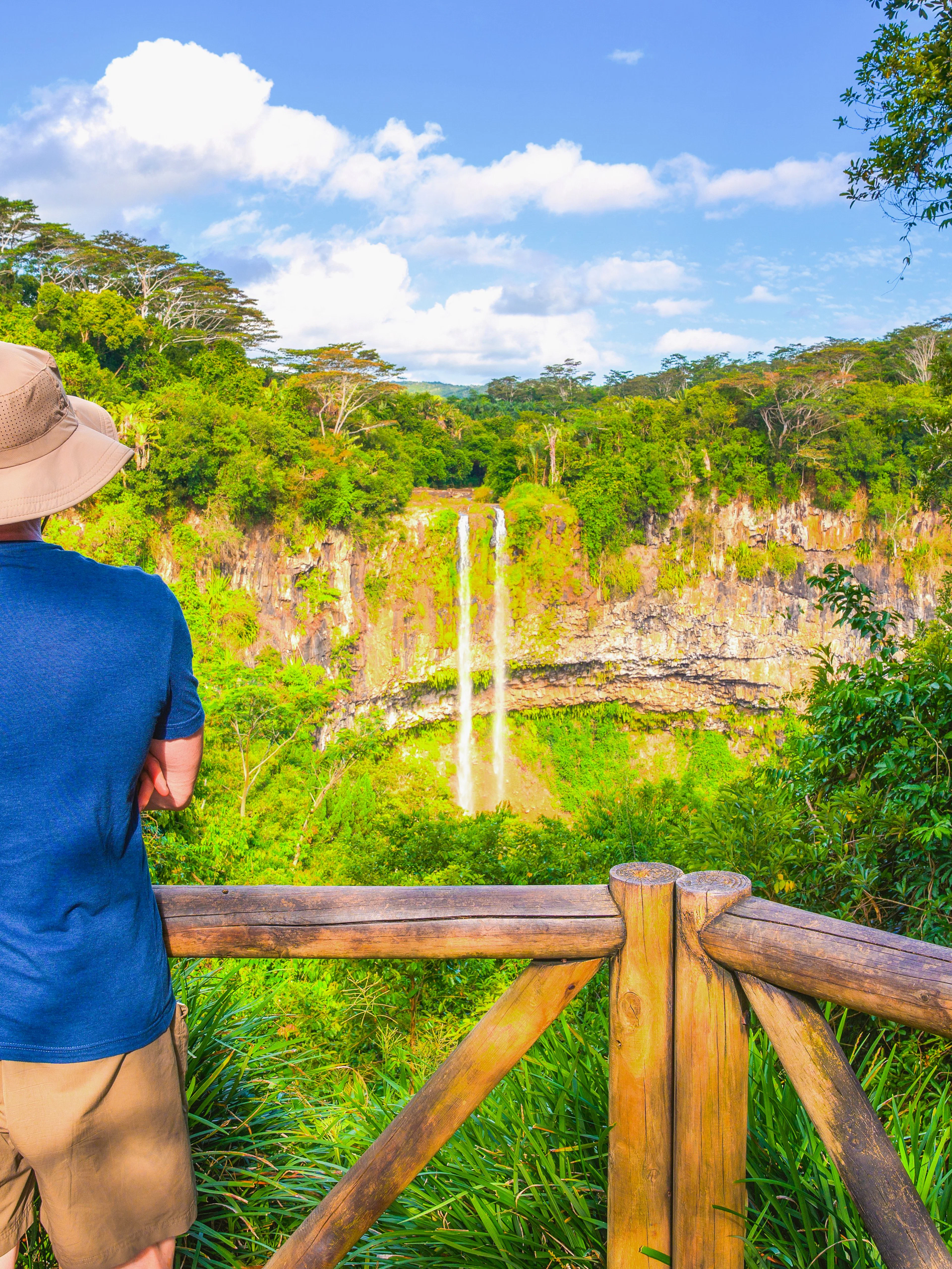 Person viewing Chamarel Waterfall from a wooden railing, surrounded by lush greenery in Mauritius.