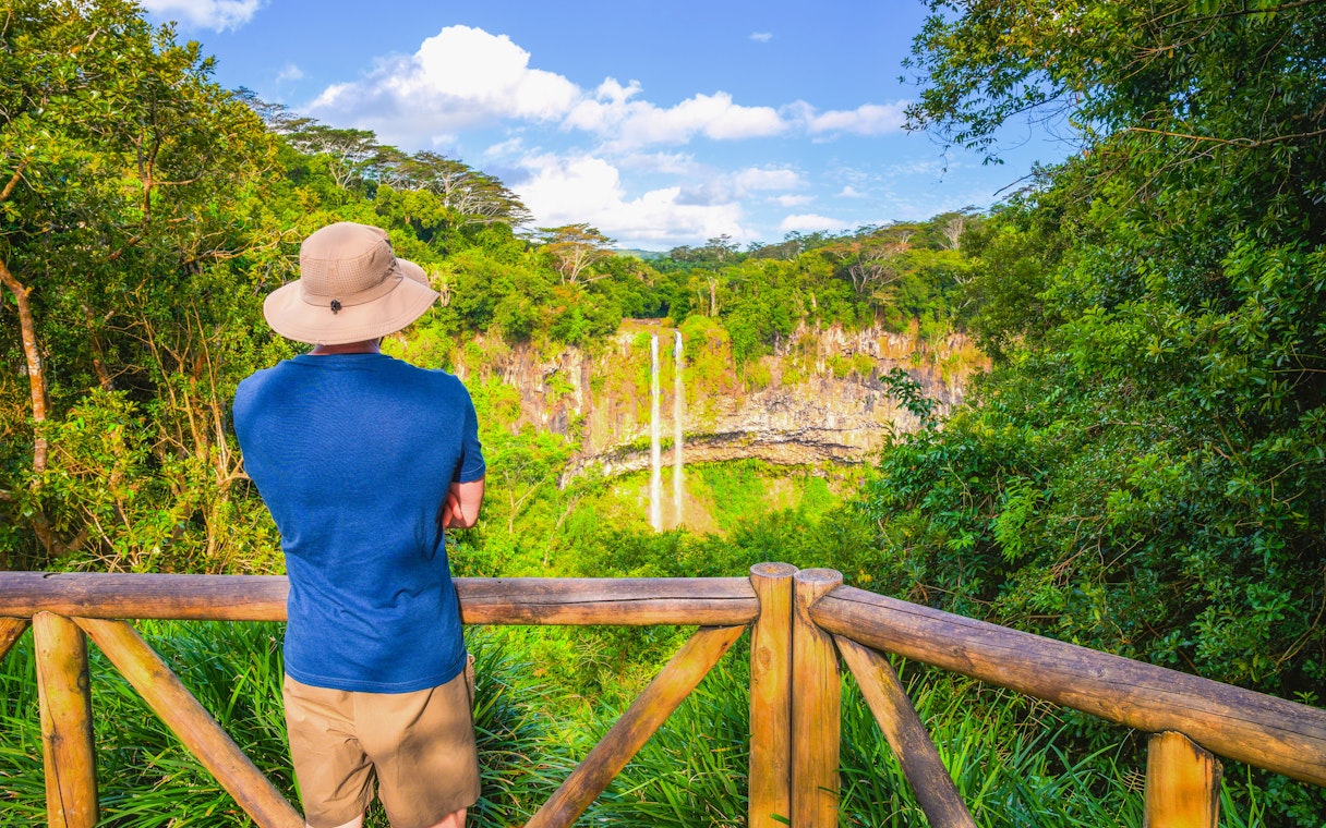 Person viewing Chamarel Waterfall from a wooden railing, surrounded by lush greenery in Mauritius.
