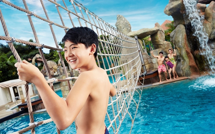 Child crossing rope bridge at Adventure Cove Waterpark, Singapore with waterfall in background.