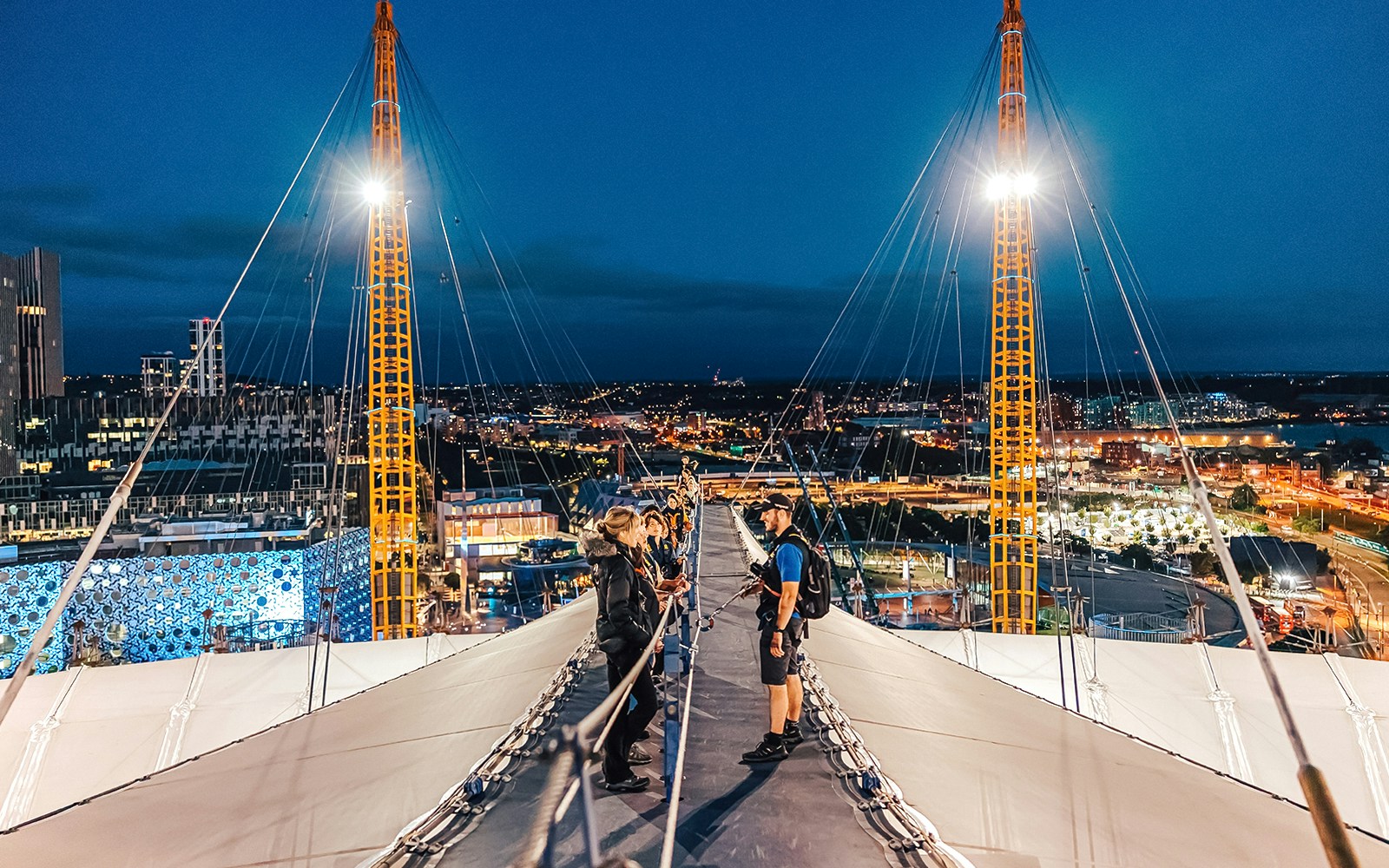 Visitors climbing Up at The O2 during twilight with city lights in the background.