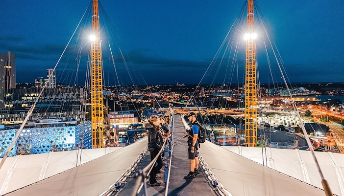 Visitors climbing Up at The O2 during twilight with city lights in the background.