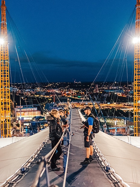 Visitors climbing Up at The O2 during twilight with city lights in the background.