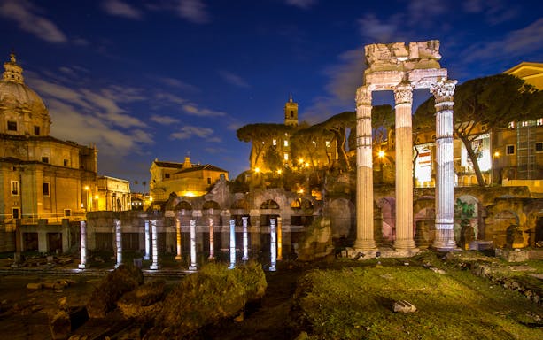 Roman Forum illuminated at night with ancient columns and surrounding architecture.