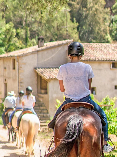Group horse riding on a trail near rustic building in countryside.