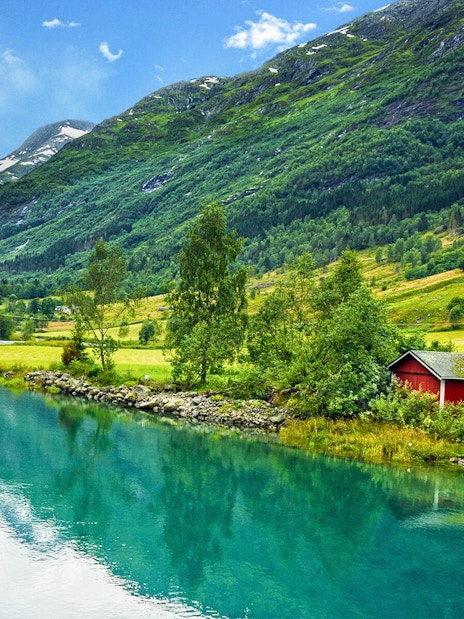 Panoramic view of a fjord with lush green mountains and a red cabin by the water.