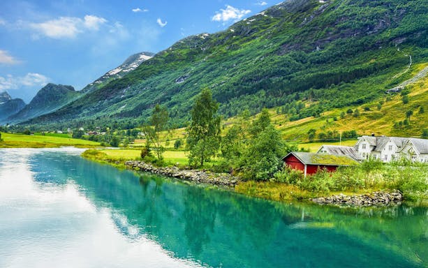Panoramic view of a fjord with lush green mountains and a red cabin by the water.