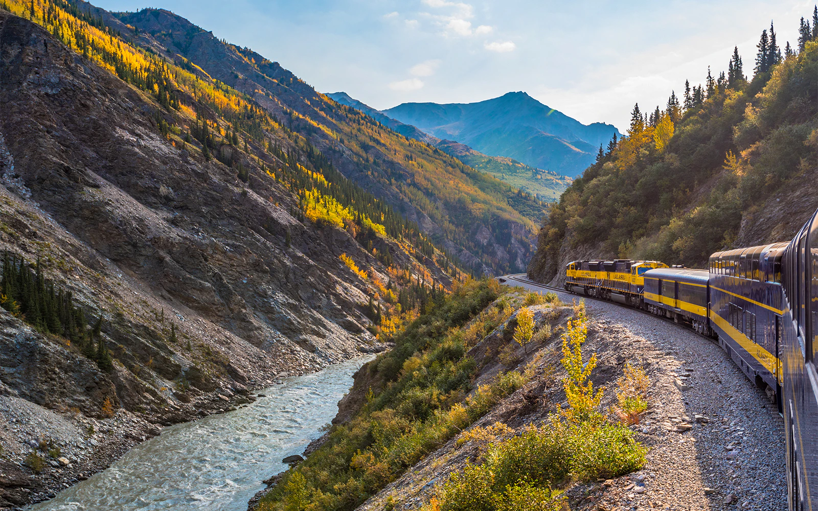 Alaska Railroad train winding through forested mountains with river below.