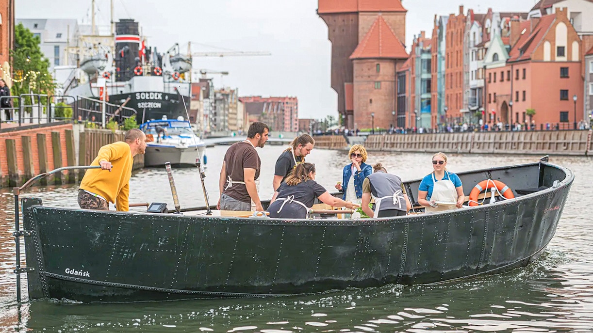 Guests tasting pierogi on a boat during Motława River cruise in Gdansk, with historic buildings in view.