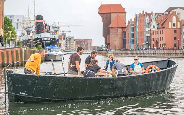 Guests tasting pierogi on a boat during Motława River cruise in Gdansk, with historic buildings in view.
