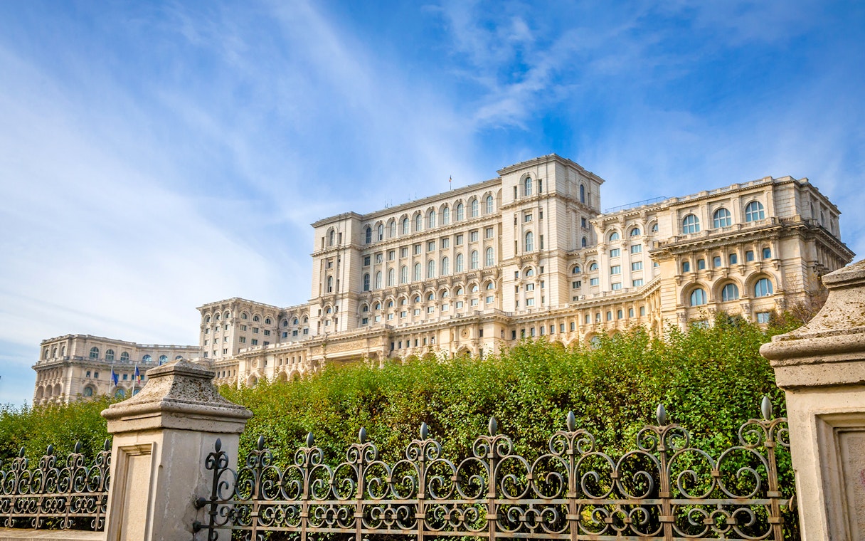 Palace of the Parliament in Bucharest, Romania, with ornate iron fence and lush greenery.
