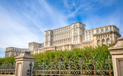 Palace of the Parliament in Bucharest, Romania, with ornate iron fence and lush greenery.