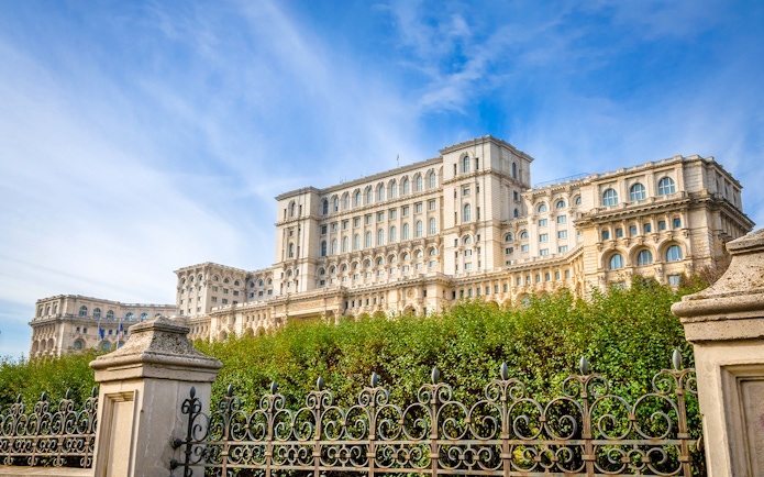Palace of the Parliament in Bucharest, Romania, with ornate iron fence and lush greenery.